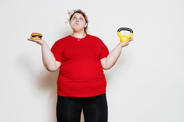 Mulher engraçada vestida de sportswear e com uma bandagem na cabeça está de pé com um hambúrguer em uma mão e o peso na outra contra uma parede branca