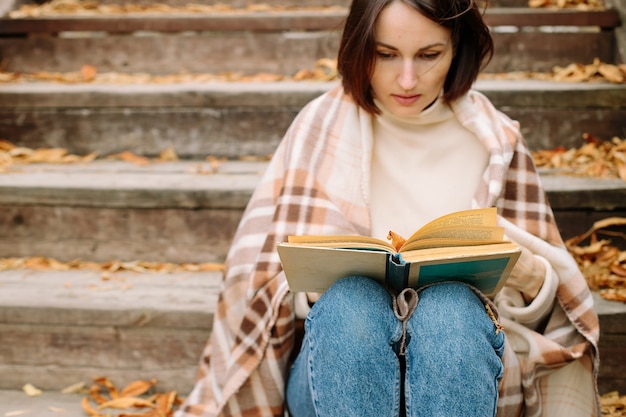 Foto mulher enrolada em um cobertor senta-se na escada e lê um livro