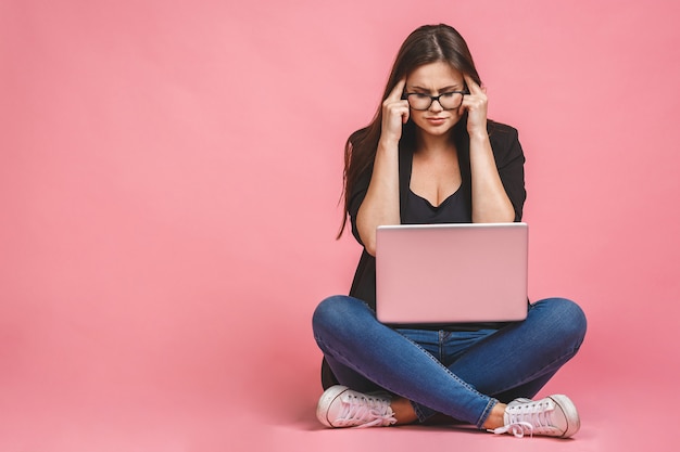 Mulher entediada triste com raiva no estresse, sentado no chão com o computador portátil isolado contra o fundo rosa.