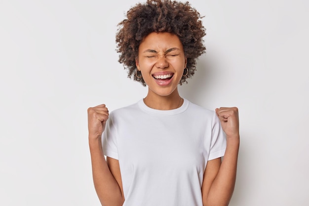 Mulher entusiasmada em êxtase com cabelo espesso encaracolado aperta os punhos e comemora o sucesso ri alegremente vestido com camiseta básica casual isolada sobre fundo branco conceito de pessoas e triunfo