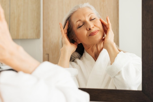 Mulher envelhecida com os olhos fechados tocando seu rosto com os dedos Fêmea em roupão fazendo massagem facial no banheirox9xA