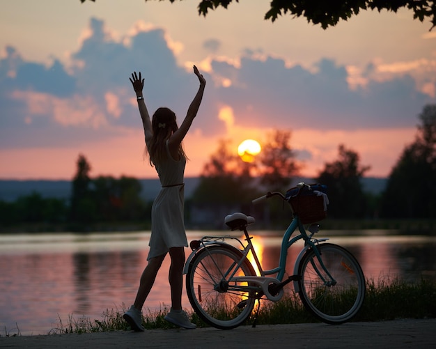 Mulher esbelta fica com os braços traseiros levantados contra o pano de fundo do pôr do sol mágico perto da bicicleta vintage