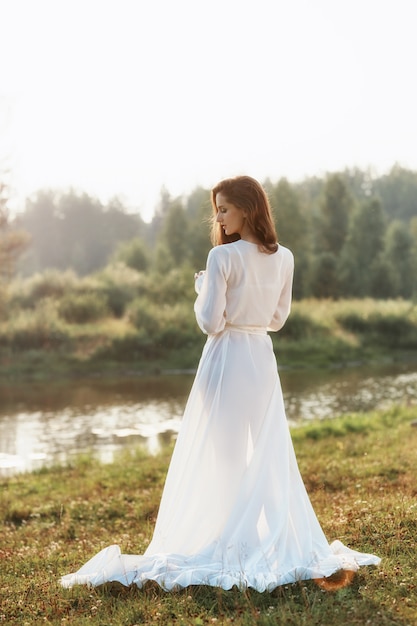 Mulher esbelta linda em um vestido branco longo caminha de manhã perto do lago.