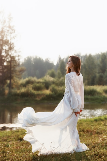 Mulher esbelta linda em um vestido branco longo caminha de manhã perto do lago.