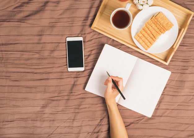 Mulher, escreve, em, grande, branca, caderno aberto, deite-se cama, com, smartphone, bandeja café da manhã