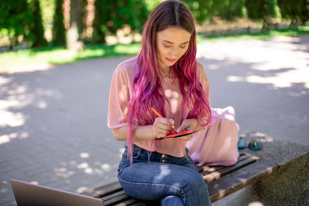 Mulher escrevendo em um caderno, sentado em um banco de madeira no parque, garota trabalhando ao ar livre em portátil.