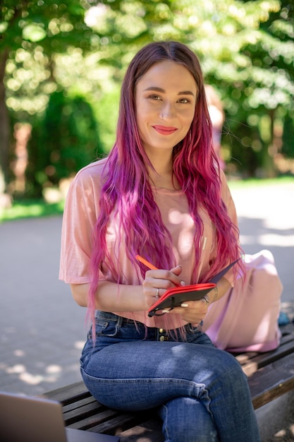 Mulher escrevendo em um caderno, sentado em um banco de madeira no parque. Garota trabalhando ao ar livre em um computador portátil, copie o espaço.