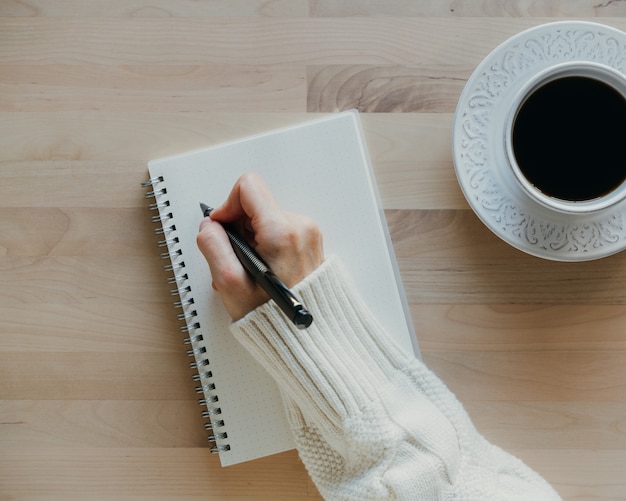 Mulher escrevendo no caderno na mesa de madeira, mão no suéter segurando caneta, desenho de caderno, vista superior