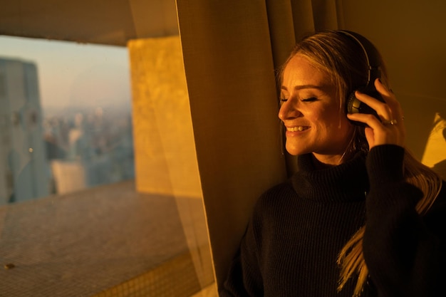 Foto mulher escuta música com fones de ouvido junto à janela com uma tecnologia de telefone móvel de vista da cidade