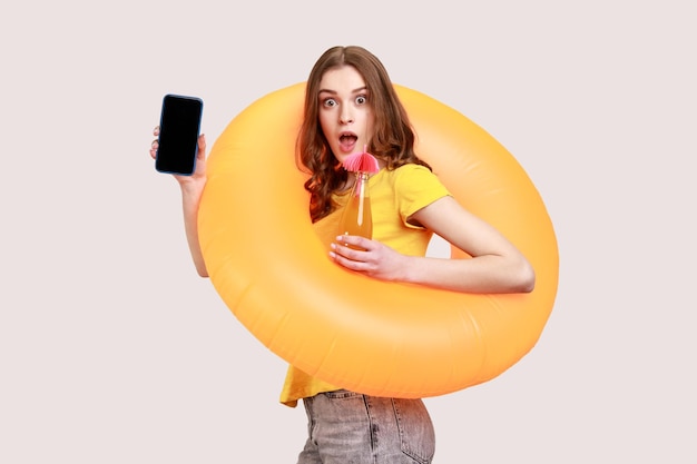 Mulher espantada de tenra idade em camiseta amarela segurando suco de anel de borracha e olhando para a câmera com expressão chocada, mostrando a tela do telefone inteligente em branco Tiro de estúdio interior isolado em fundo cinza