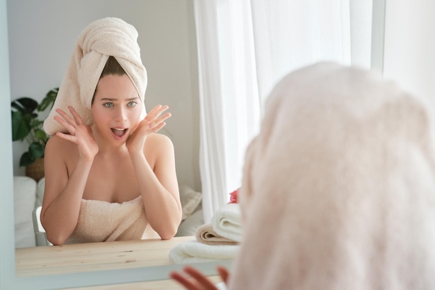 Mulher espantada envolta em toalhas olhando para o reflexo do espelho com a boca aberta na mesa durante a rotina diária na sala de luz