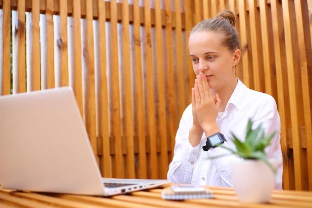 Mulher esperançosa vestindo camisa branca sentada no café ao ar livre trabalhando no laptop esperando por e-mails importantes rezando por boa sorte educando online