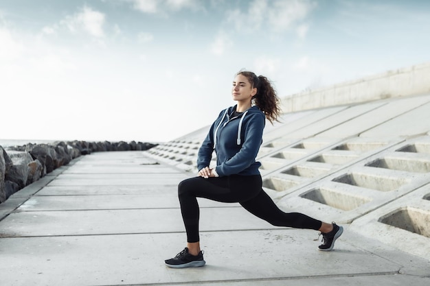 Mulher esportiva encaracolada em roupas esportivas fazendo aquecimento corporal antes do treino na praia urbana Estilo de vida saudável