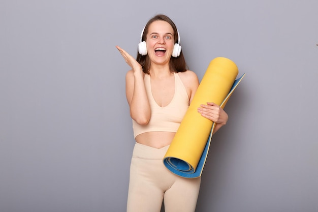 Mulher esportiva feliz vestida com roupas esportivas bege carrega karemat enrolado para prática de ioga desfruta de bom som em fones de ouvido isolados sobre fundo cinza