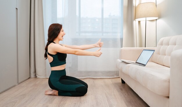 Mulher esportiva fica feliz em fazer ioga online com um laptop na sala de estar do housefitness e ...