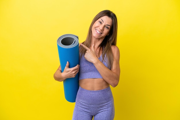 Mulher esportiva indo para aulas de ioga enquanto segura um tapete isolado de fundo não amarelo apontando para o lado para apresentar um produto