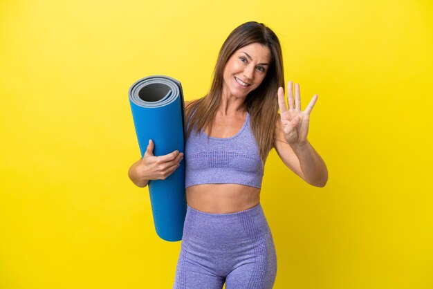 Mulher esportiva indo para aulas de ioga enquanto segura um tapete isolado não amarelo feliz e contando quatro com os dedos