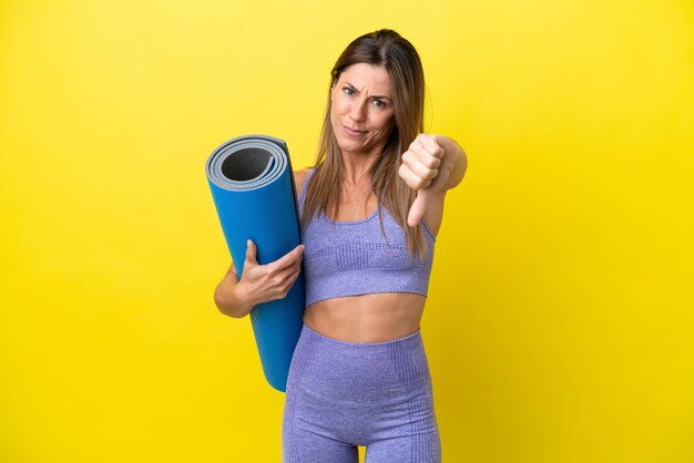 Mulher esportiva indo para aulas de ioga enquanto segura um tapete isolado não amarelo mostrando o polegar para baixo com expressão negativa