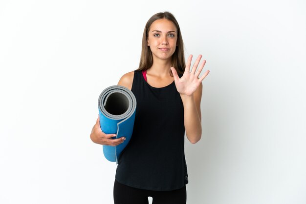 Mulher esportiva indo para aulas de ioga segurando um tapete sobre um fundo branco isolado, contando cinco com os dedos