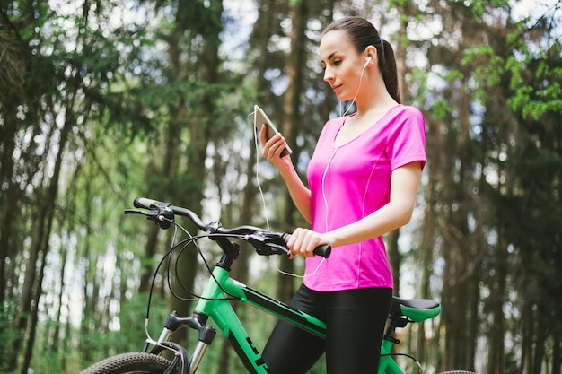 Mulher esportiva na bicicleta Mountait na floresta enquanto andava com o telefone na mão Apps para Bike Sport