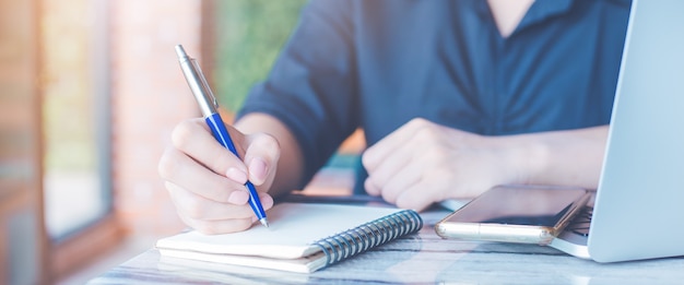 Mulher está escrevendo em um caderno com uma caneta no escritório. Na mesa, há telefones celulares e computadores portáteis. Banner da Web.