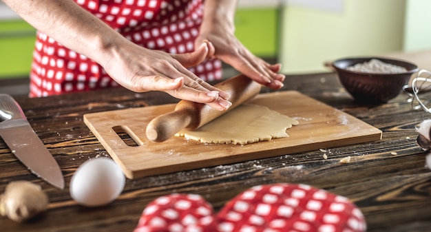 Mulher está estendendo massa crua em uma mesa de madeira com um rolo para fazer deliciosos biscoitos caseiros em sua cozinha