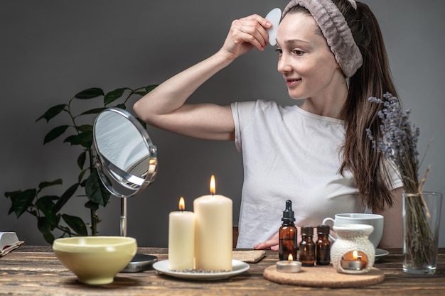 Mulher está olhando no espelho e fazendo uma massagem com um raspador de guache velas e uma lâmpada de aroma com óleos essenciais na mesa conceito de cuidados faciais aromaterapia rituais matinais e noturnos