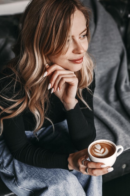 Mulher está olhando para longe e ela está sentada em um sofá e segurando uma xícara de café.