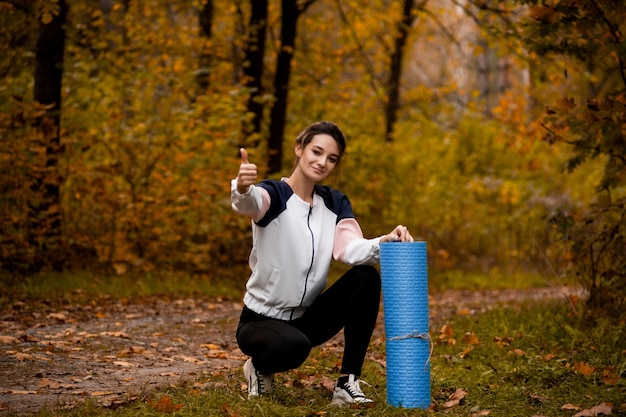 Mulher está satisfeita com seu treino. Floresta ou parque na forma traseira. Jovem apta a mulher latejando e sentada perto de seu tapete de ioga. Floresta de outono. Praticando ioga. Alongamento antes de correr.