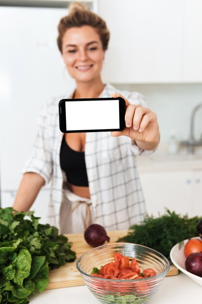 Mulher está segurando smartphone com tela em branco para seu projeto durante o cozimento na cozinha moderna
