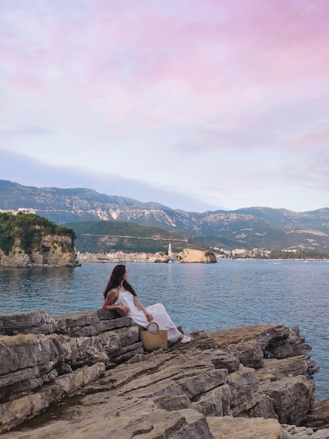 Mulher está sentada em uma rocha com a cidade velha e montanhas em Budva Montenegro