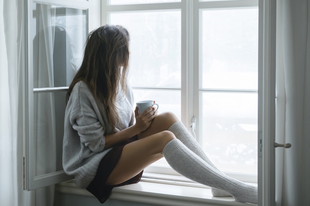 Mulher está sentada no parapeito da janela e bebendo chá ou café quente