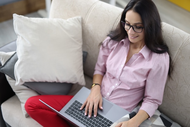 Mulher está sentada no sofá e trabalhando no laptop.