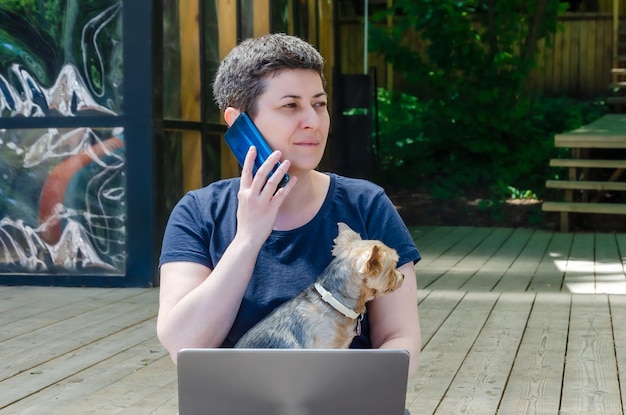 Mulher está trabalhando ao ar livre no laptop, segurando o cachorro Yorkshire terrier em seus braços, falando ao telefone