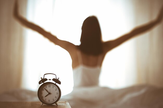 Foto mulher estendendo as mãos na cama depois de acordar, fulgura de sol. brunette entrando em um dia feliz e relaxado depois de uma boa noite de sono e vista de trás. conceito de um novo dia e fim de semana alegre.