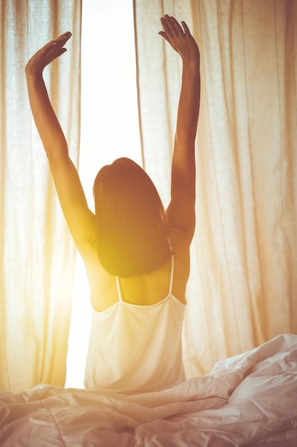 Mulher esticando as mãos na cama depois de acordar, reflexo do sol. Morena entrando em um dia feliz e relaxado após uma boa noite de sono e vista traseira. Conceito de um novo dia e fim de semana alegre.