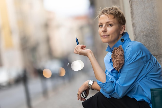 Mulher estilosa adulta com cabelo curto sentada na rua europeia e fumando um cigarro eletrônico