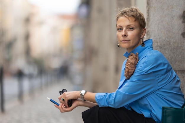 Mulher estilosa adulta com cabelo curto sentada na rua europeia e segurando um cigarro eletrônico