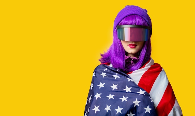 Mulher estilosa com óculos de realidade virtual e agasalho esportivo dos anos 80 com a bandeira dos eua na parede amarela