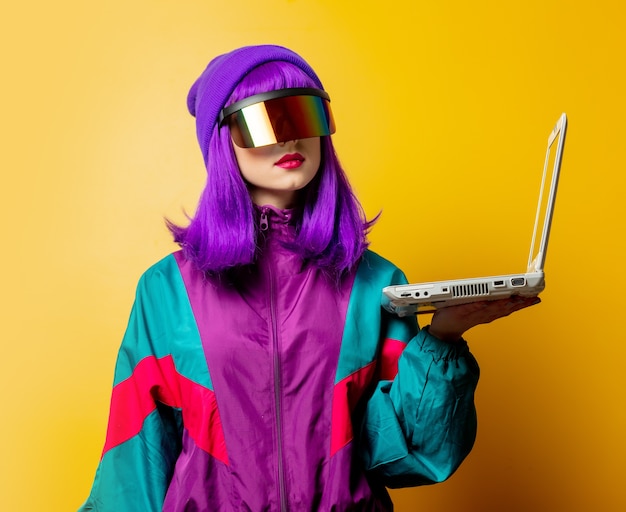 Mulher estilosa com óculos de realidade virtual e agasalho esportivo dos anos 80 com laptop na parede amarela