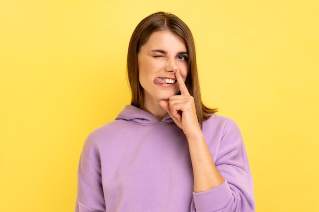 Mulher estranha louca com cabelo castanho, segurando o dedo no nariz, mostrando a língua menina entediada inculta se divertindo com maus modos vestindo capuz roxo Tiro de estúdio interior isolado em fundo amarelo