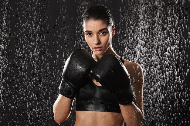 Mulher estrita fazendo esportes e boxe em luvas em pé na posição de defesa sob gotas de chuva, isoladas sobre fundo preto