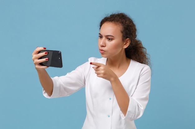 Mulher estritamente médica afro-americana isolada em fundo azul. Médico em bata médica fazendo selfie filmado no celular fazendo videochamada. Conceito de medicina pessoal de saúde. Simule o espaço da cópia.