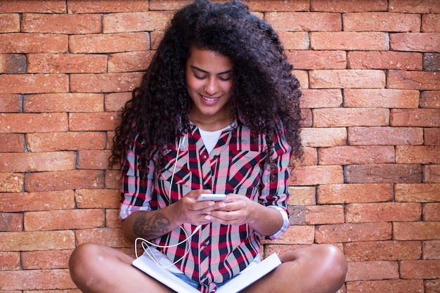 Mulher estudante feliz com penteado afro, sentado com o fundo da parede de tijolos, usando telefone celular e sorrindo.