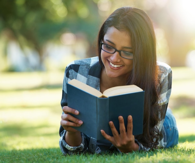 Mulher estudante feliz e lendo livro na grama verde para estudar literatura ou história na natureza Pessoa feminina inteligente ou jovem adulto com sorriso ou óculos para aprendizagem de capítulos ou educação no parque