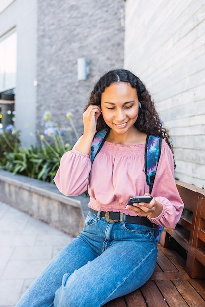 Mulher estudante universitária latina sorrindo e usando seu celular sentado fora do shopping em tempo livre