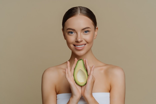 Mulher europeia calma e sorridente com cabelo penteado segura o abacate como recomendação para uma máscara facial nutritiva