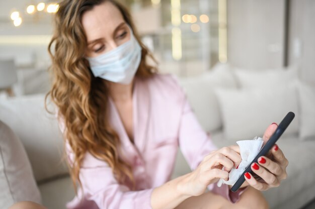 Mulher européia com máscara facial, limpando o telefone com desinfetante para as mãos, usando algodão com álcool para limpar e evitar contaminação com o vírus corona. limpeza de celular para eliminar germes, covid-19.