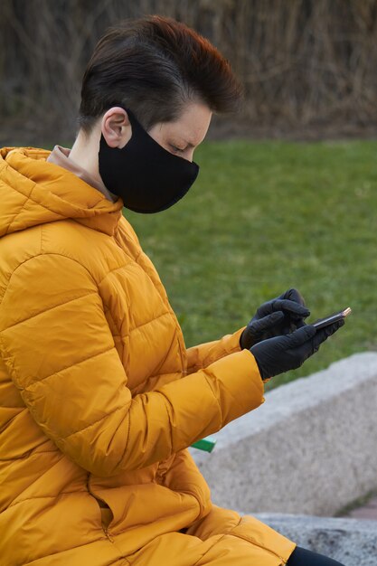 Mulher européia de meia idade em máscara protetora preta e luvas segurando um smartphone fora durante o coronavírus epidemia de covid-19