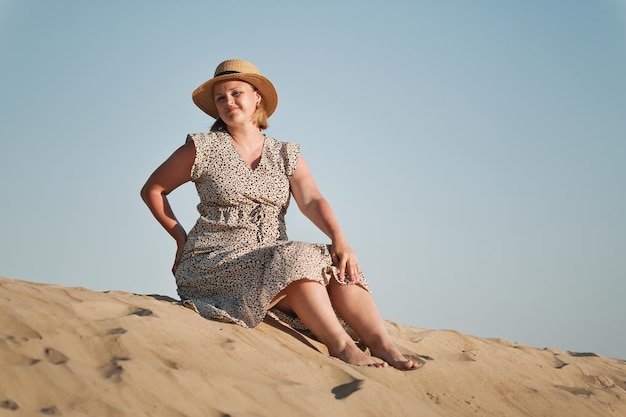 Mulher europeia feliz com vestido longo e chapéu de palha senta-se na areia e desfruta de férias no deserto